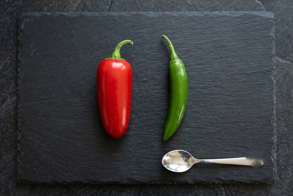 Fresh red Jalapeño pod beside fresh green Serrano pod on matte slate with silver teaspoon for scale