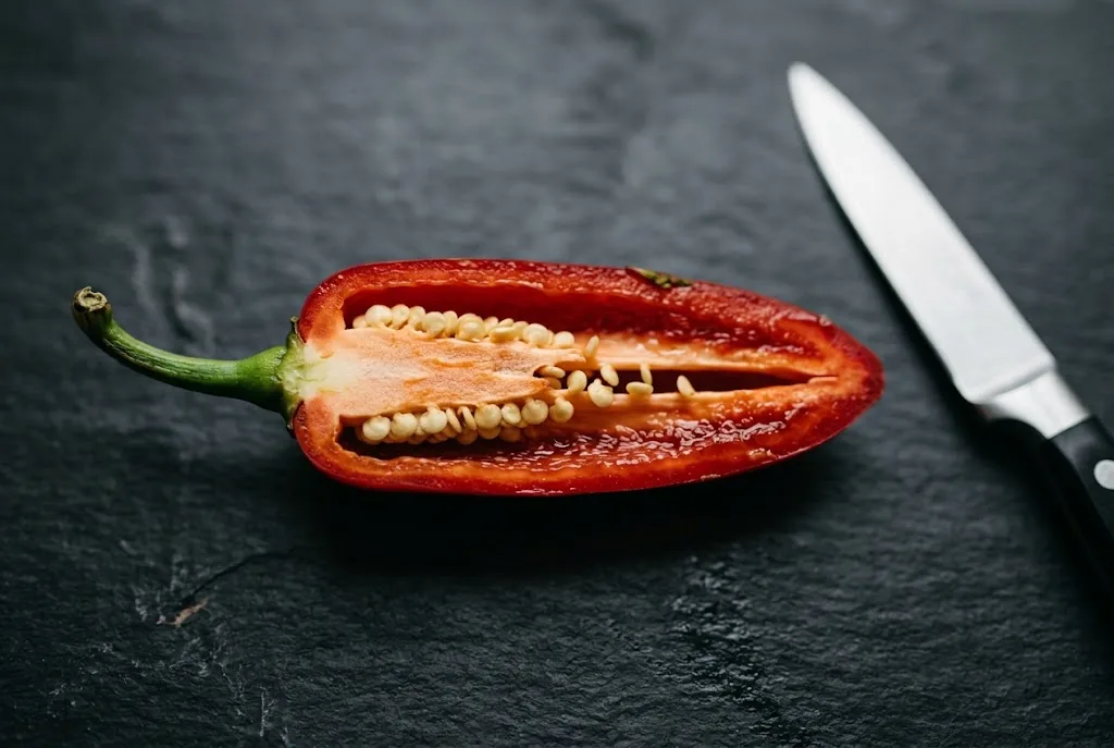 A halved fresh red ripe jalapeño pepper laid open on dark matte slate showing the pale cream pith and yellow seeds inside the cavity, chef's knife resting a few inches to the right